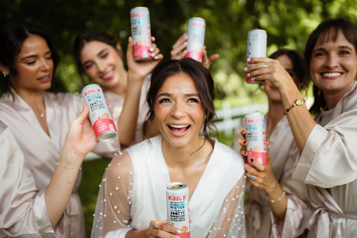 Bride and bridesmaids in robes joyfully holding canned drinks aloft during wedding morning preparations.
