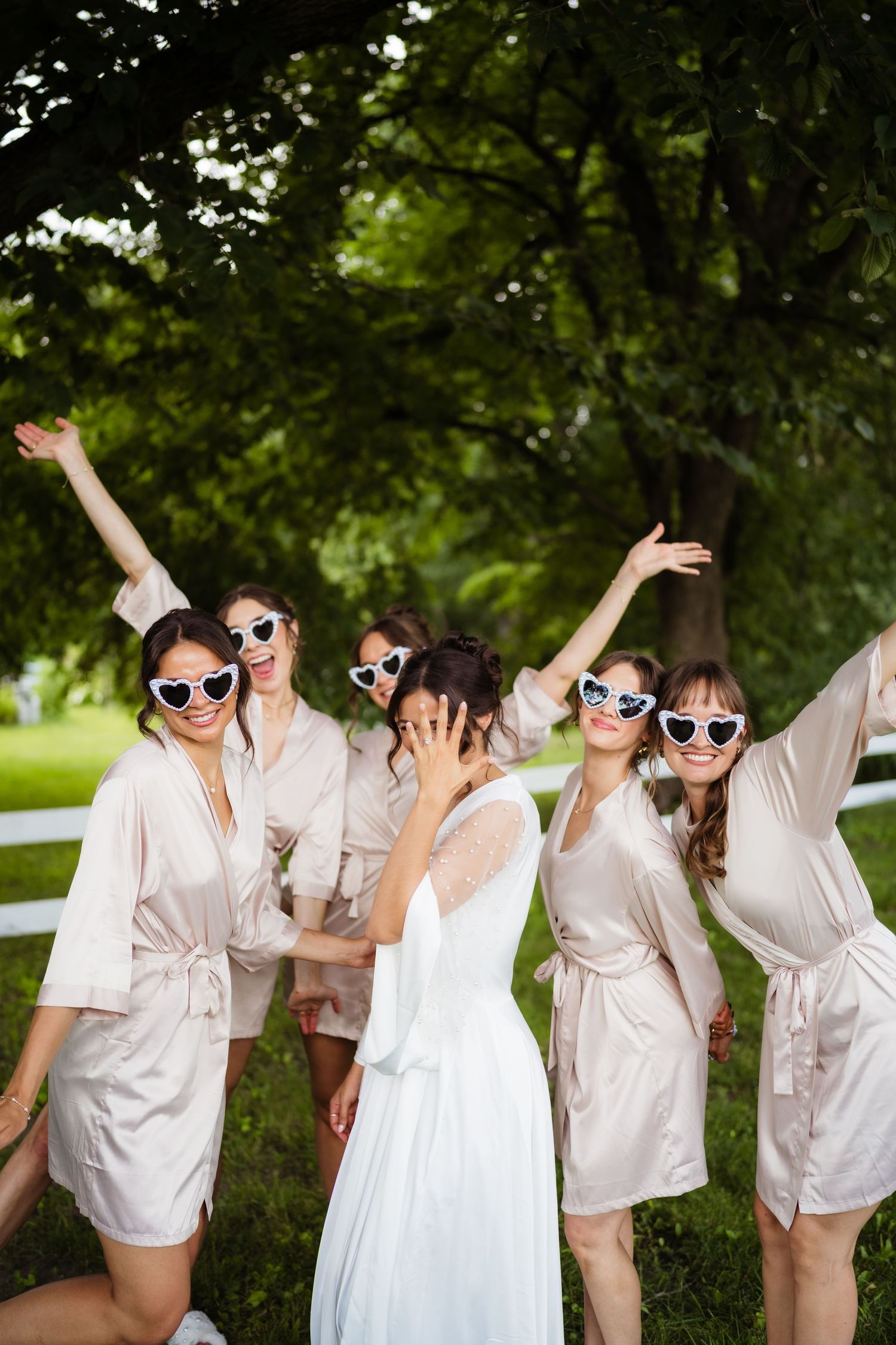 Bride in white gown with bridesmaids in champagne robes and heart sunglasses celebrating outdoors.