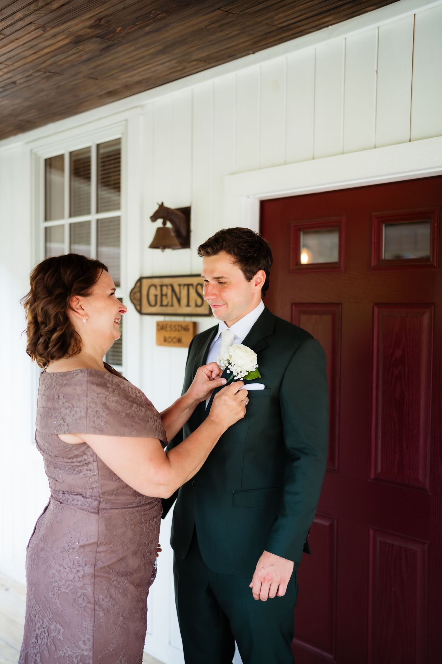 A woman in a mauve lace dress pins a boutonniere on a groom in a dark green suit outside a rustic venue.