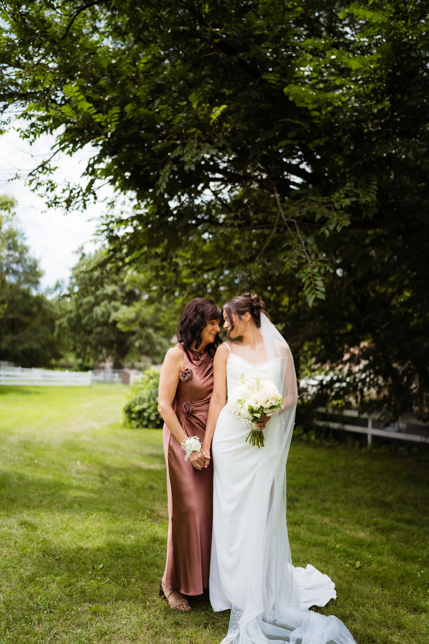 Bride in white gown holding bouquet stands with bridesmaid in mauve dress, sharing an intimate moment outdoors under lush green trees.