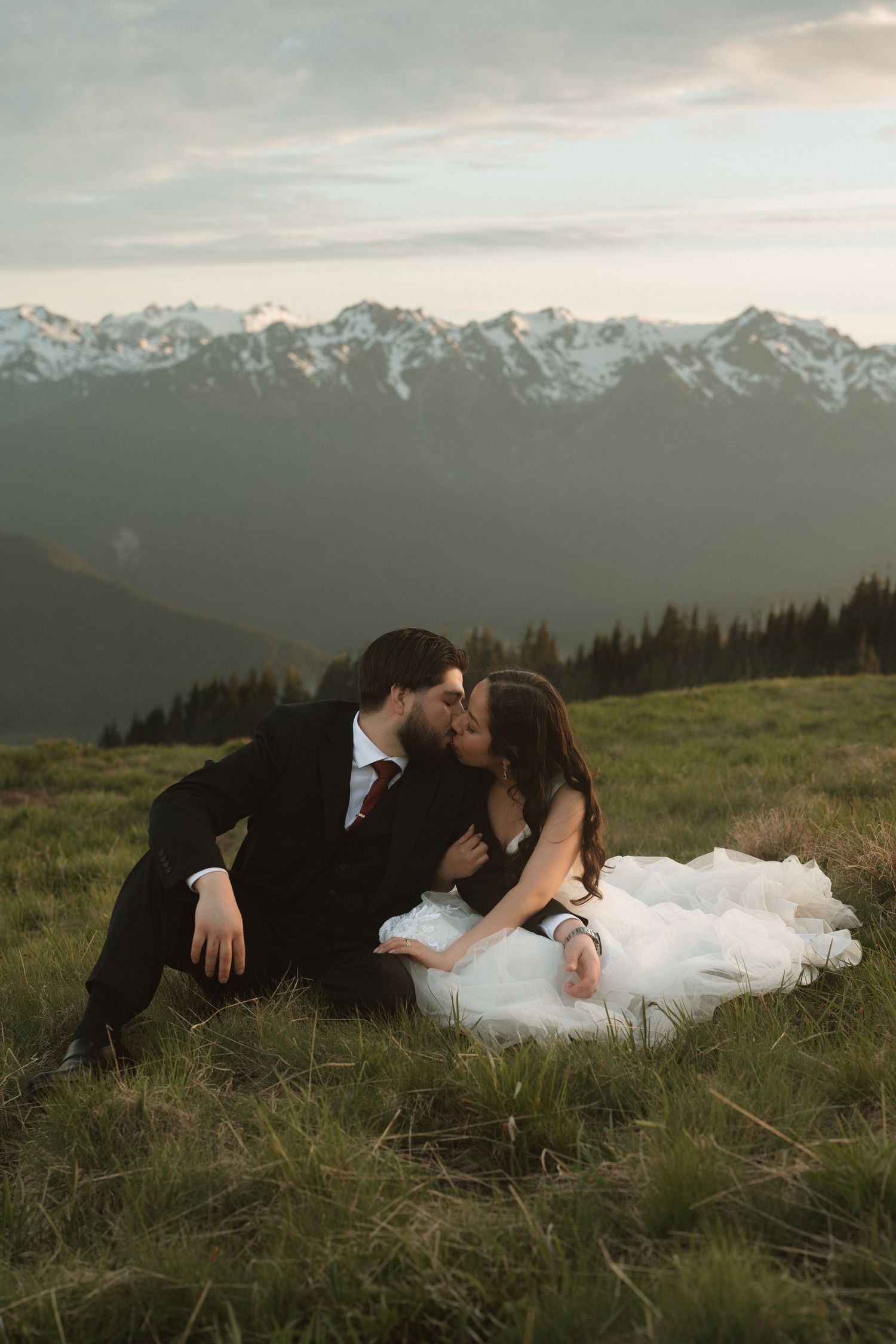 Bride and groom share a romantic kiss on a grassy mountain meadow with snow-capped peaks in the background.