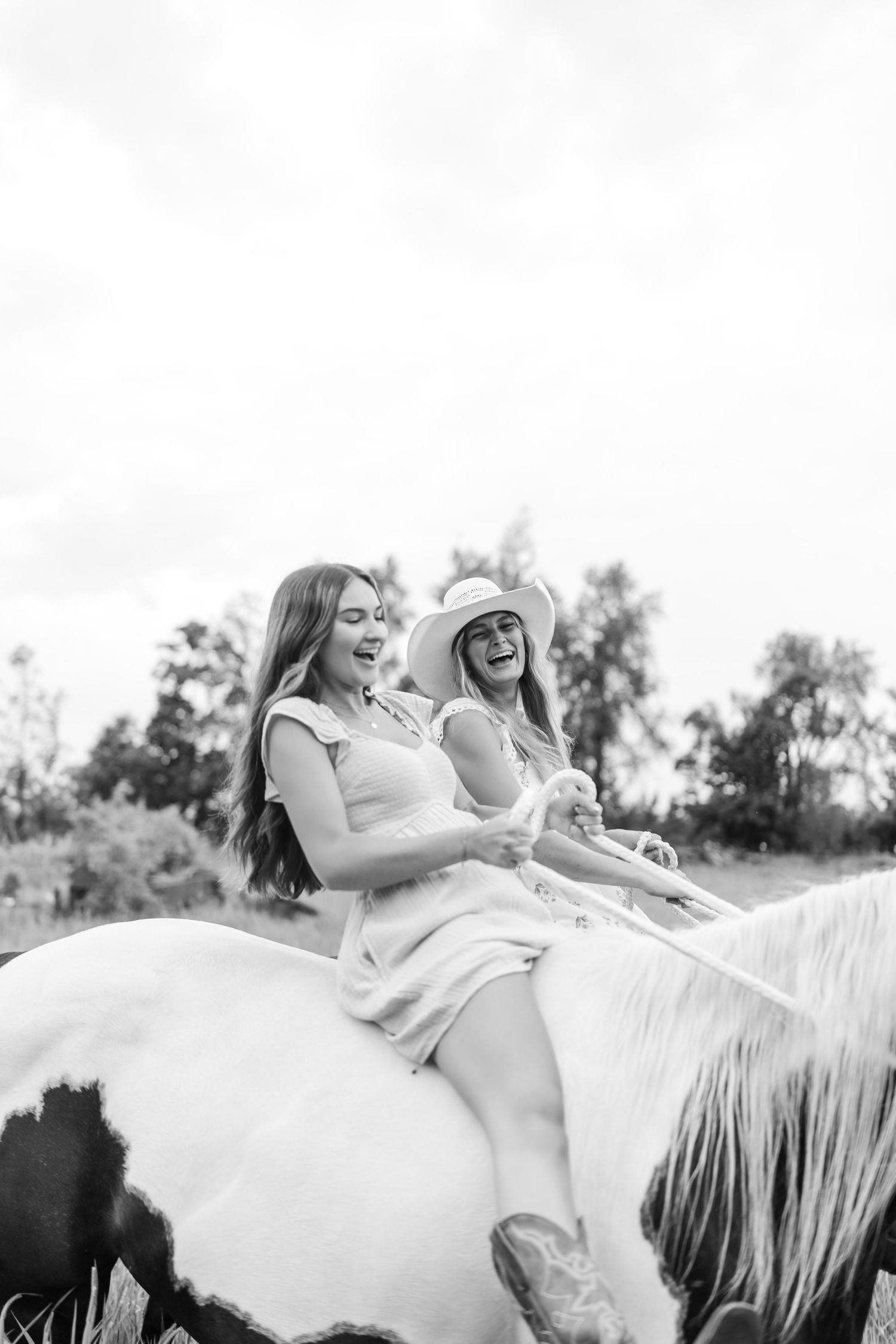 Two laughing women ride a pinto horse together in a sunny field, one wearing a cowboy hat, in black and white photo.