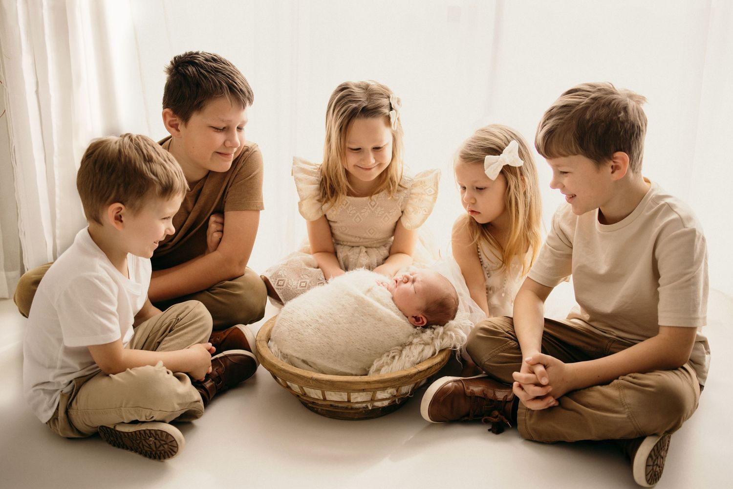 Five children sitting together on a white floor, lovingly gathered around a sleeping newborn baby in a wicker basket.