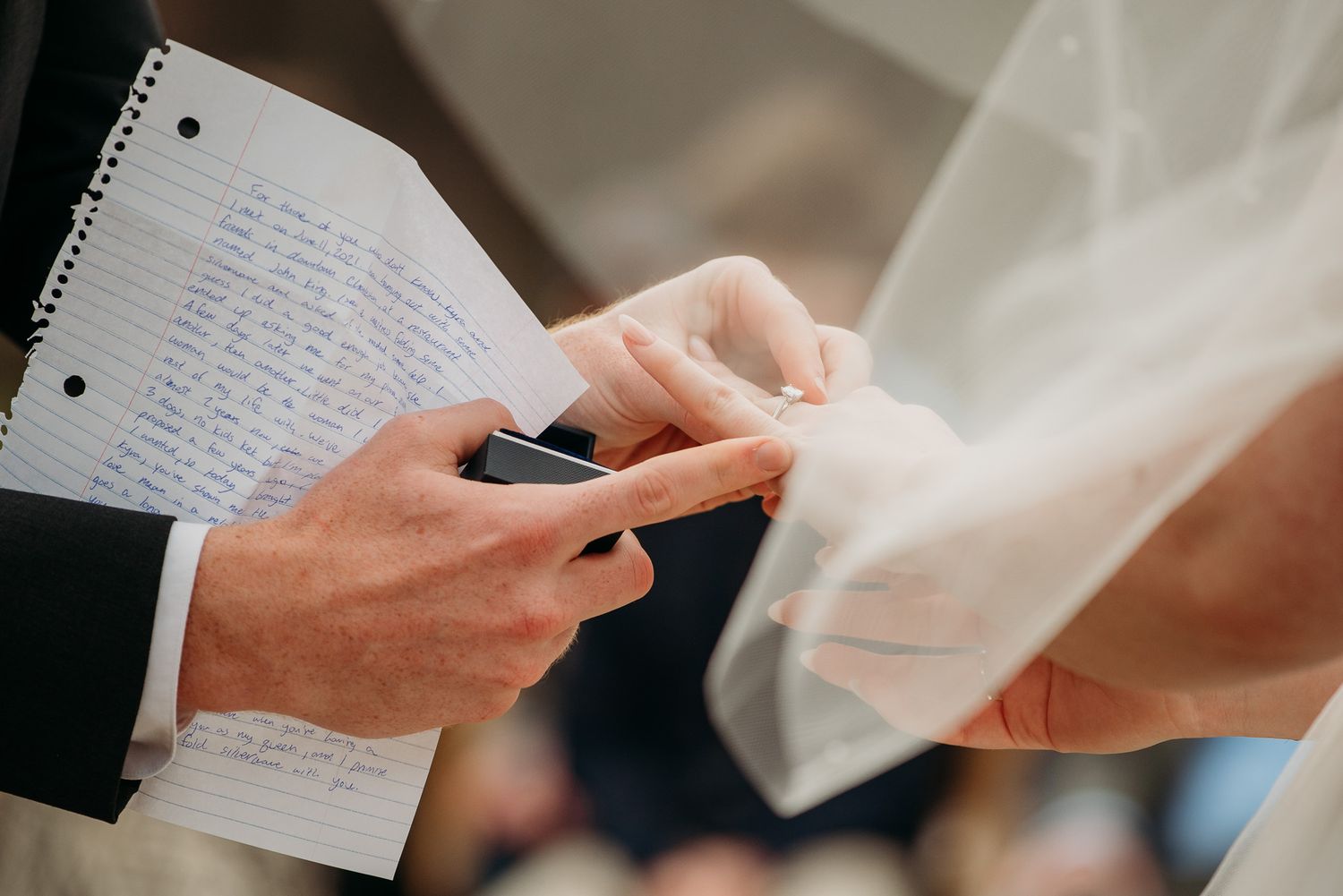 Close-up of groom placing wedding ring on bride's finger during ceremony, with handwritten vows visible.