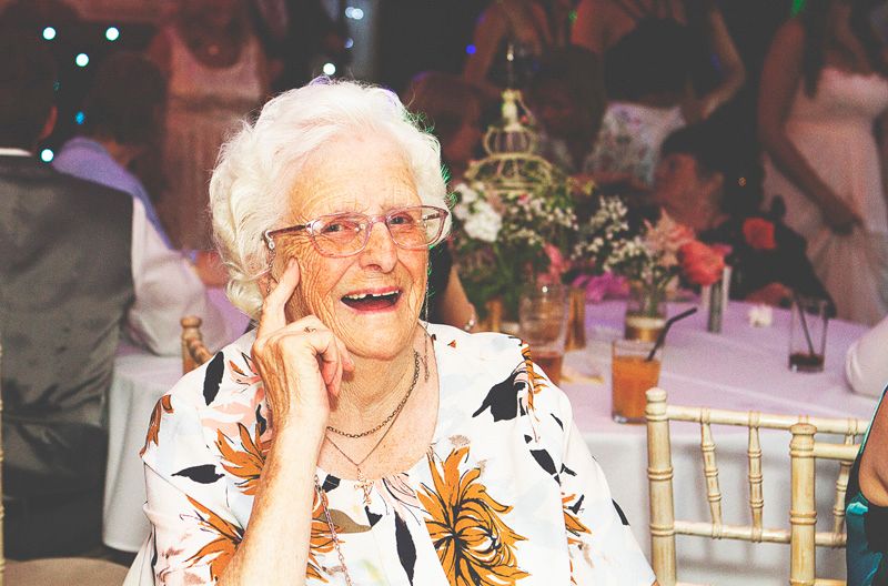 Elderly woman laughing while talking on phone at a festive event, wearing floral blouse and glasses.