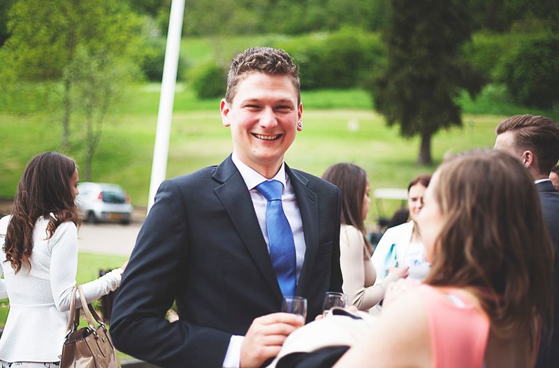 Smiling man in navy suit and blue tie holding a drink at an outdoor social gathering event.