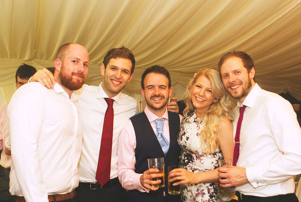 Five smiling friends pose together at a wedding reception, dressed in white shirts and ties, holding drinks.
