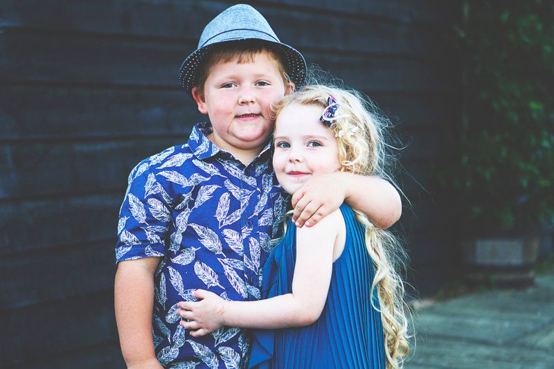 Two young children, a boy in a blue feather-print shirt and hat hugging a girl in a blue dress with blonde hair.