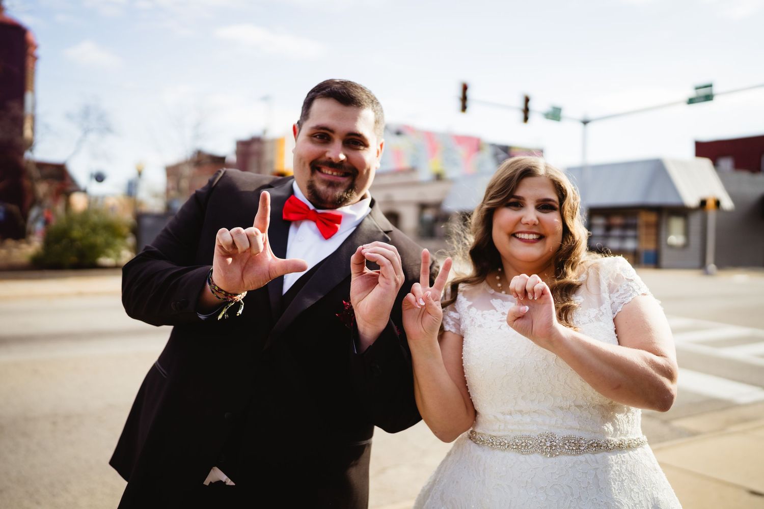 Bride and groom smiling and making hand gestures on a city street corner, groom in tuxedo with red bow tie.