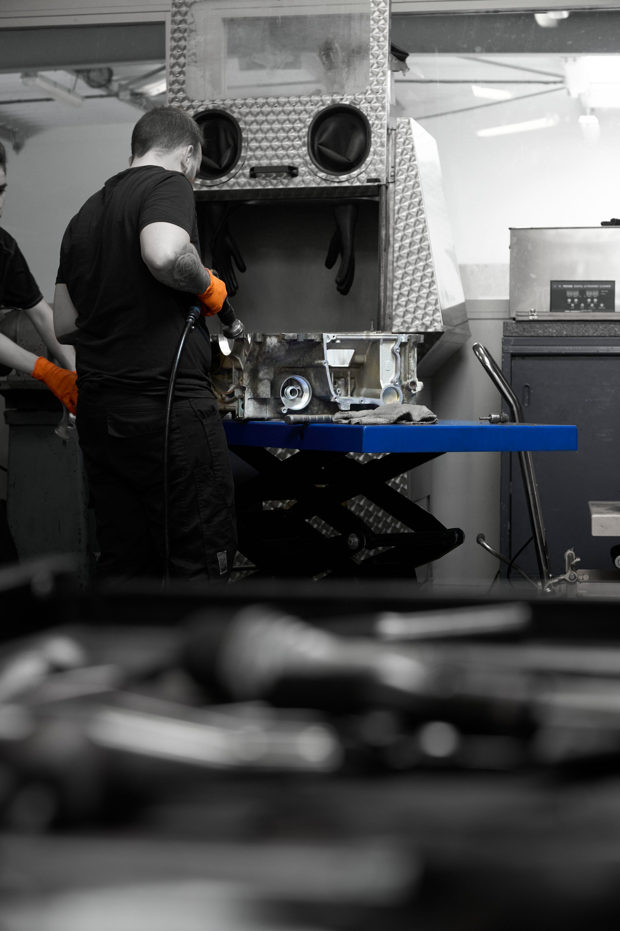 Mechanic in black uniform with orange gloves working on engine component in professional auto repair shop.
