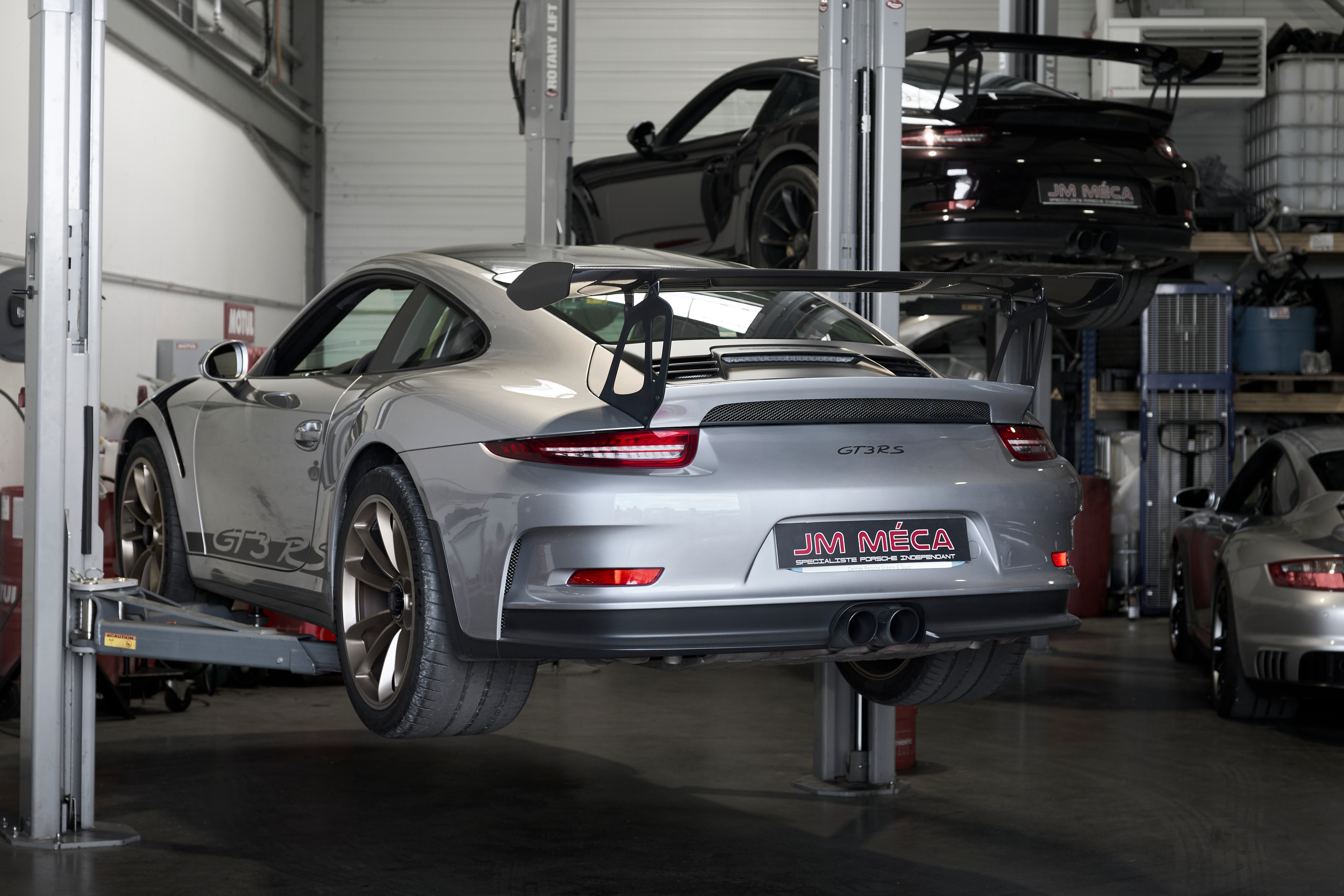 Silver Porsche 911 GT3 RS rear view on hydraulic lift inside professional sports car service garage.