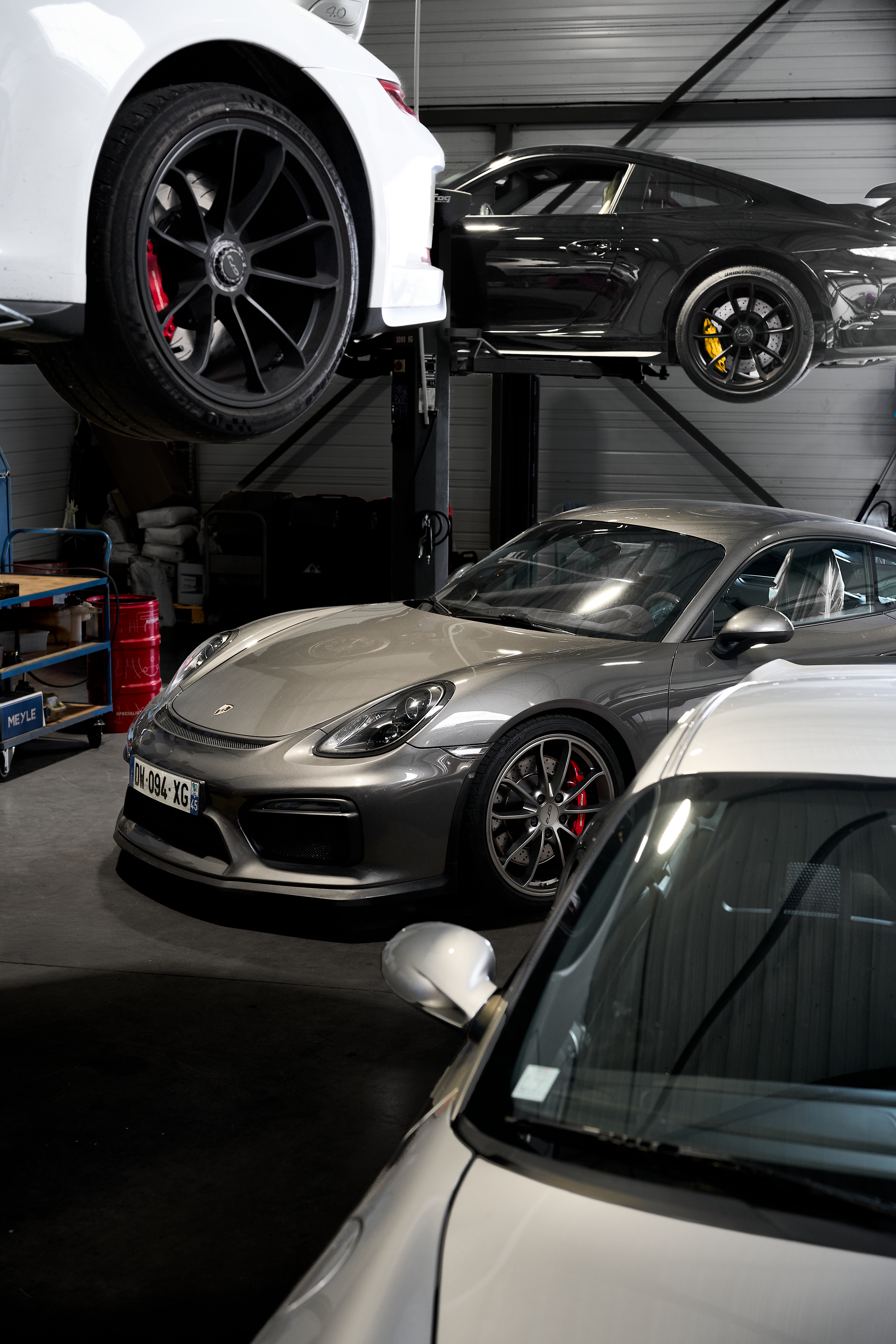 Multiple Porsche sports cars including Cayman GT4 stacked on lifts inside a specialized repair garage.