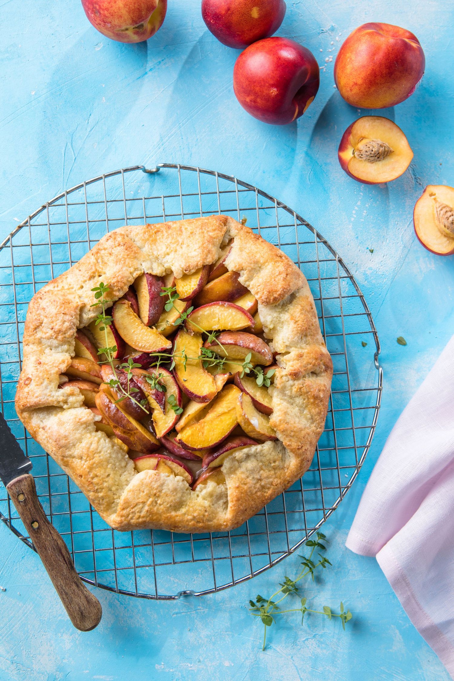 Rustic peach galette on a cooling rack with fresh nectarines on a blue background.