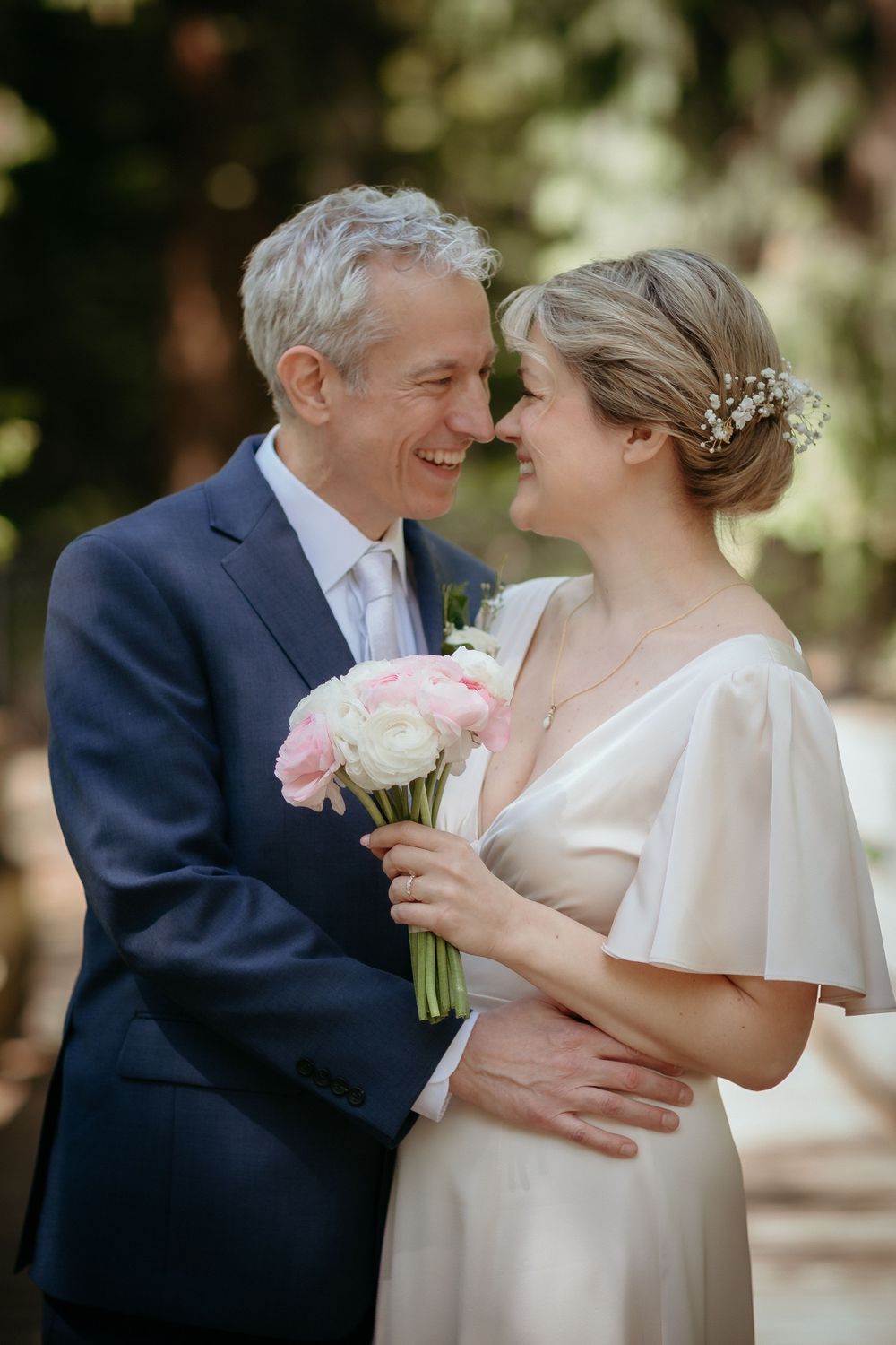 A bride and groom smile at each other, she holds pink roses, wearing a white gown with floral hair accessory.