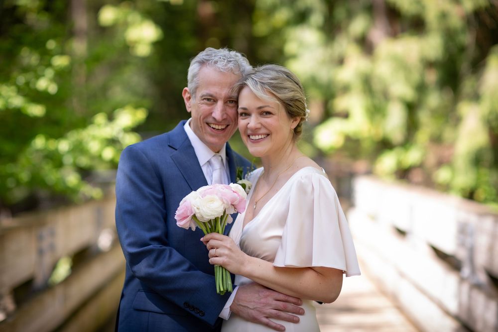 Happy couple in wedding attire smiling on wooden bridge surrounded by lush green trees, bride holding pink roses bouquet.