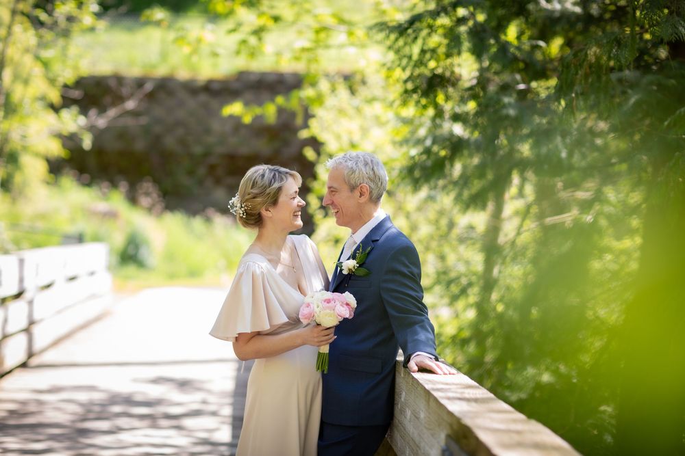 Couple sharing a romantic moment on a wooden bridge surrounded by lush greenery, bride holding pink flowers.
