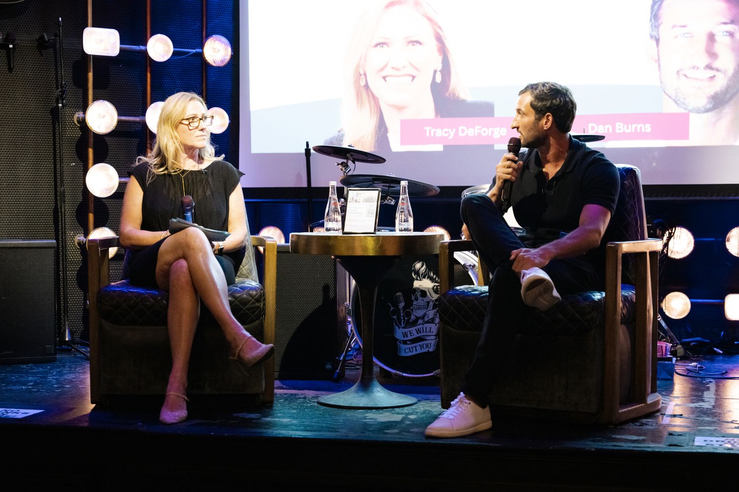 Two speakers engage in a panel discussion on a staged venue with bright lights and a presentation screen behind them.