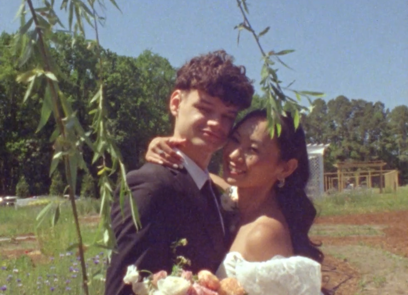 A smiling couple in wedding attire poses under a willow tree outdoors on a sunny day.