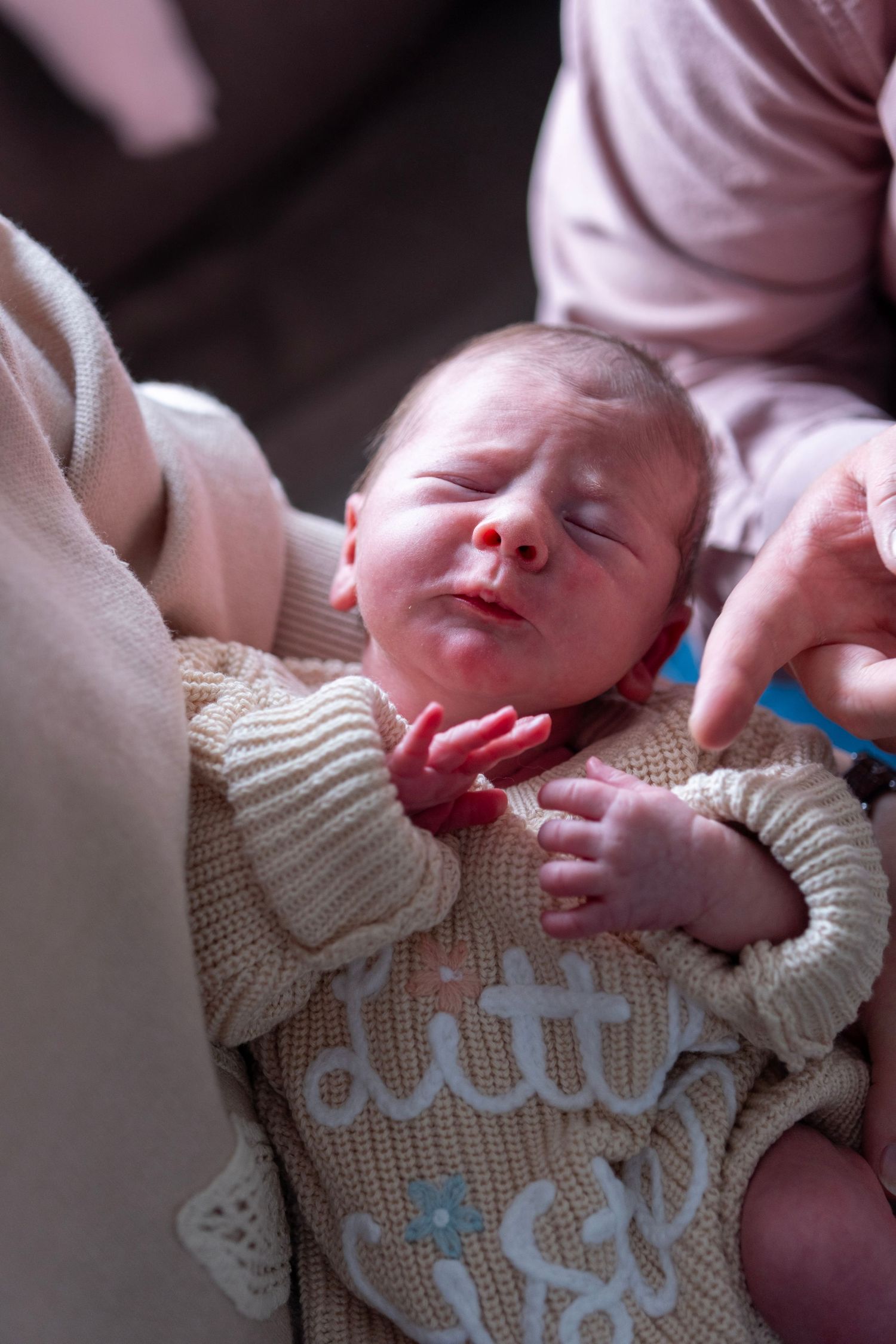 Newborn baby sleeping peacefully in a knit onesie reading 'little sis', cradled in a caregiver's arms.