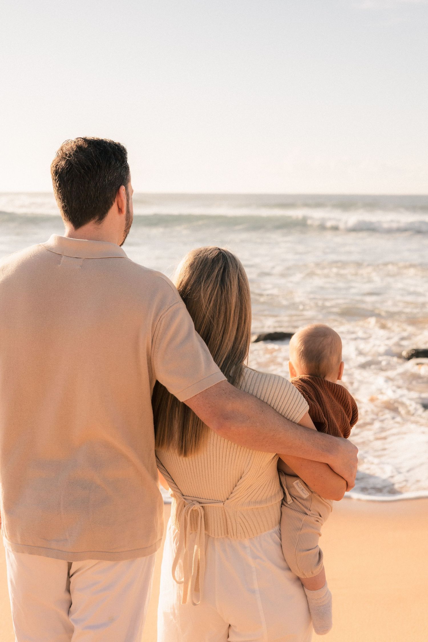 Family of three standing together on a sandy beach, parents embracing their baby while gazing at ocean waves at sunset.