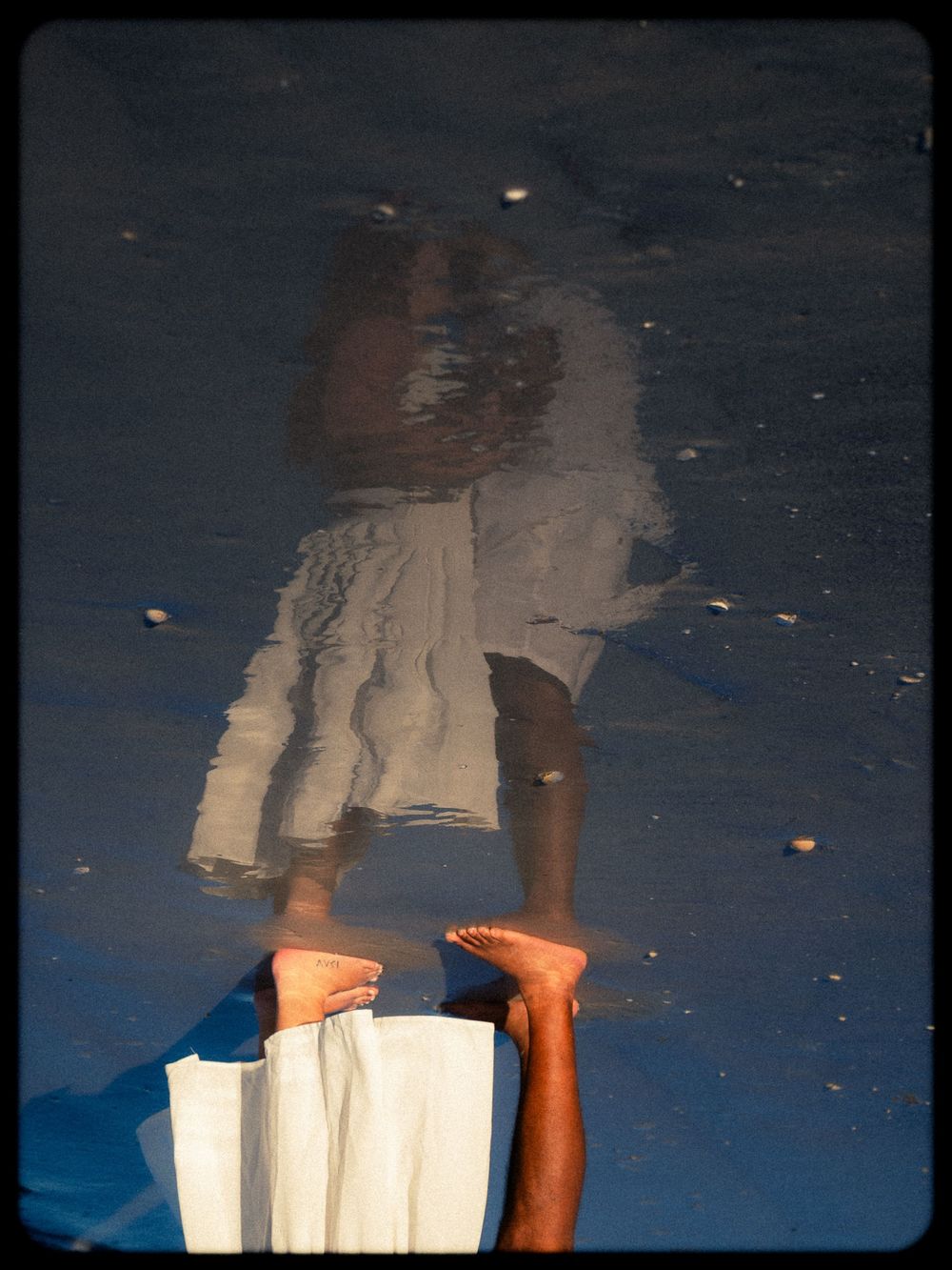 Reflection of a person in white skirt on wet beach sand with bare feet visible at bottom of frame.