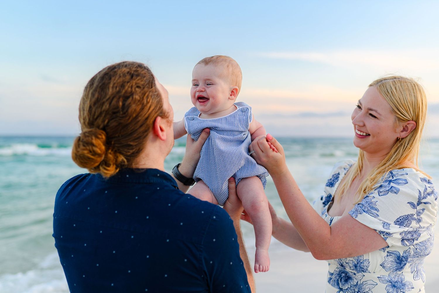 Parents lift their smiling baby at sunset on a beautiful beach with turquoise ocean waves behind them.