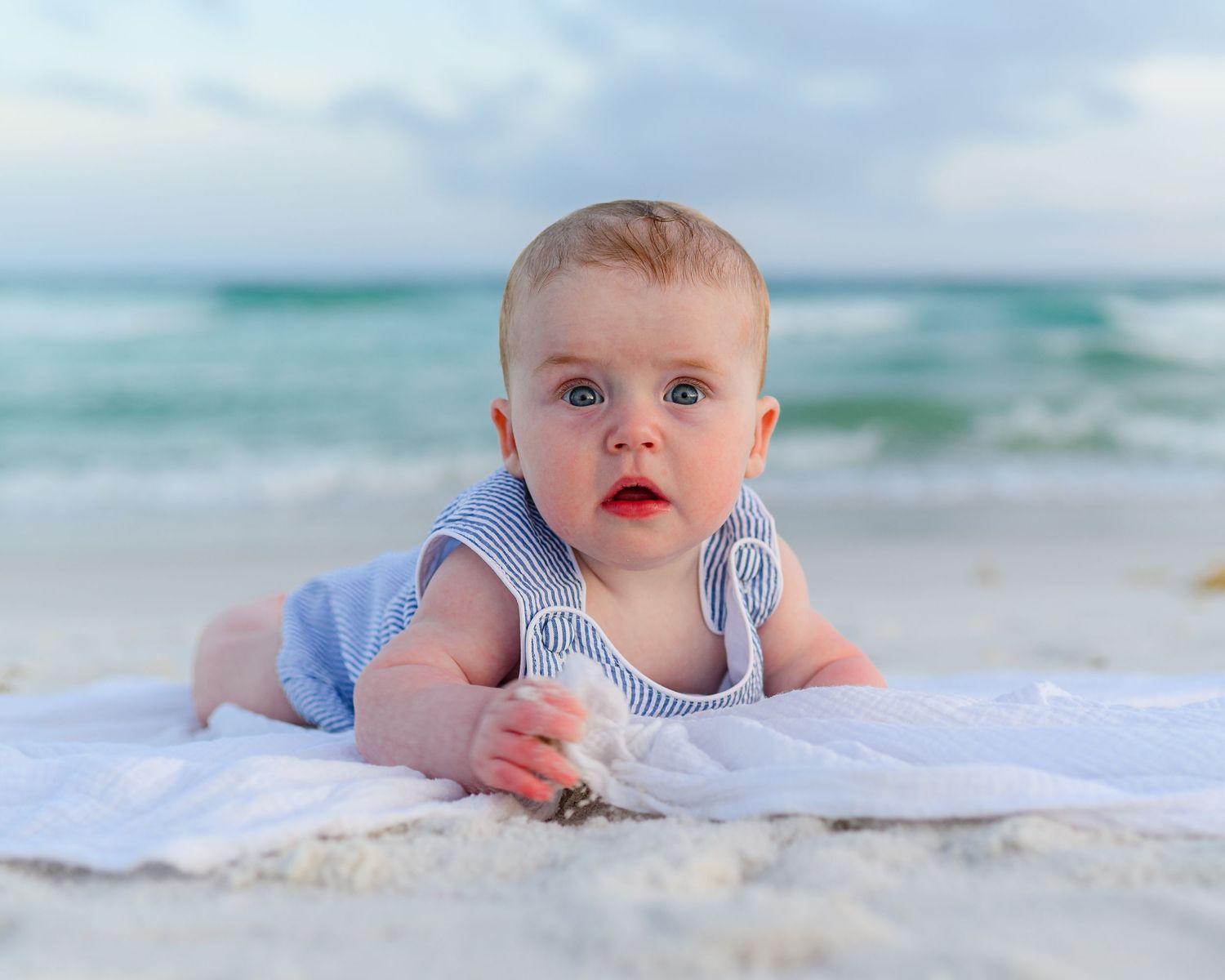 A cute baby in a blue romper lies on a white blanket on a sandy beach with ocean waves in the background.