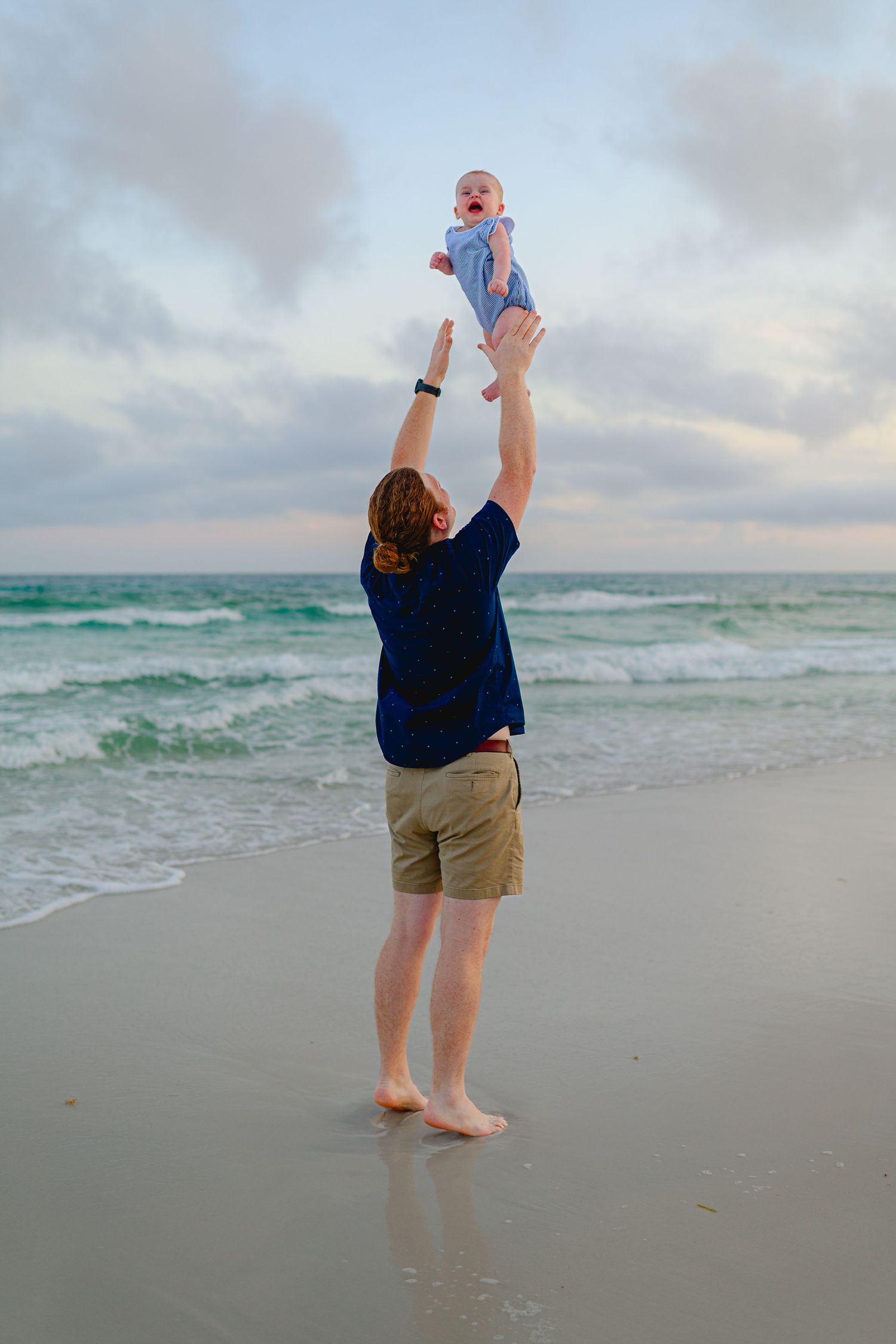 A father joyfully lifts his baby high in the air on a scenic beach with turquoise waves and a cloudy sky.