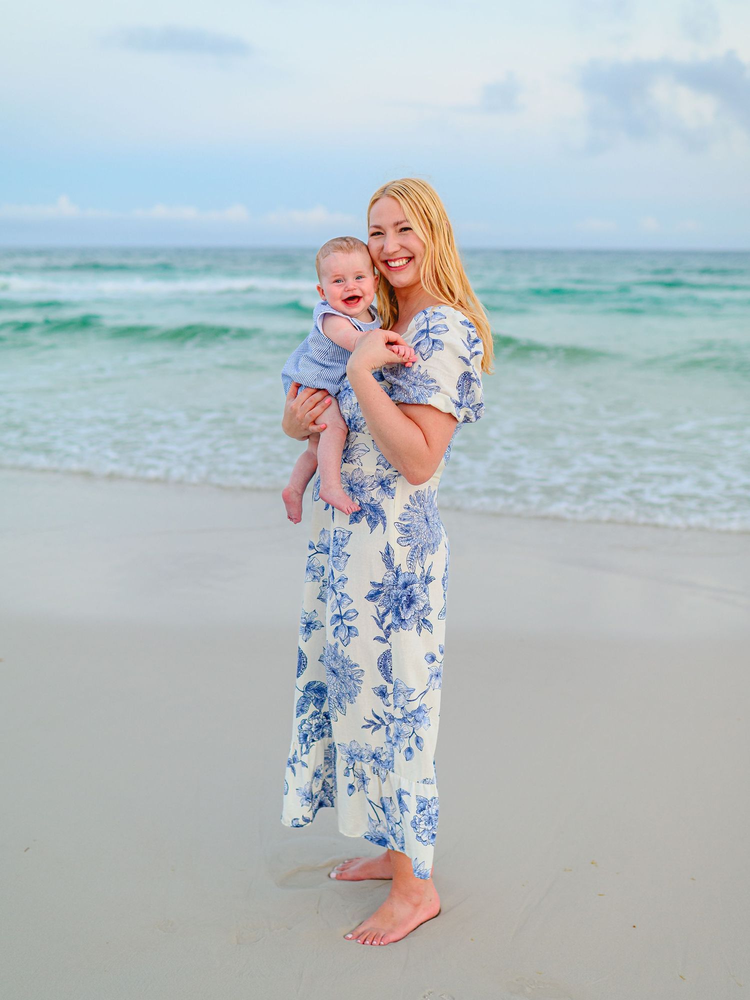 A smiling blonde woman in a blue floral dress holds a baby on a sunny beach with turquoise waves behind them.