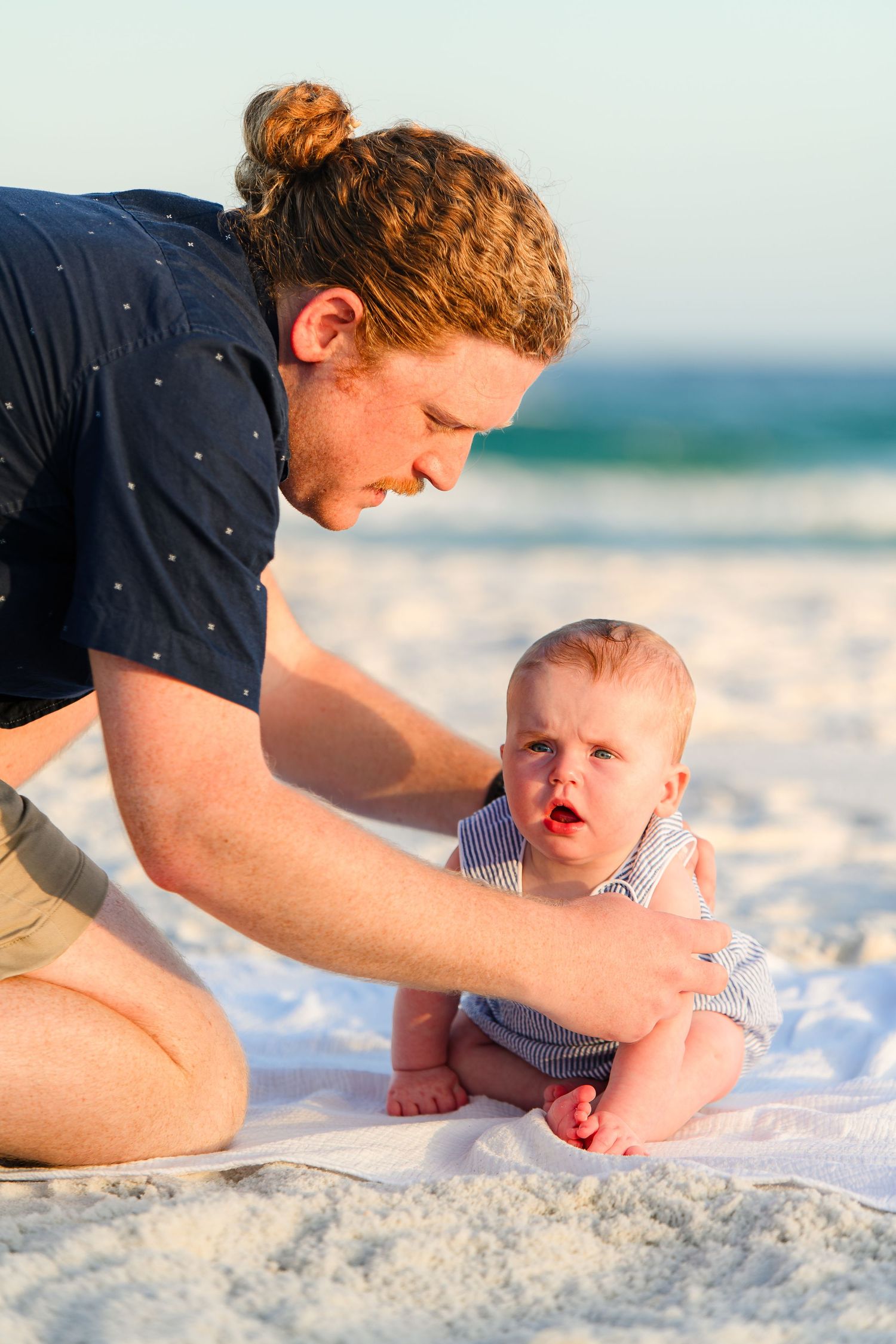 A red-haired father kneels on white sand beach, tenderly supporting his baby boy in a striped outfit at sunset.