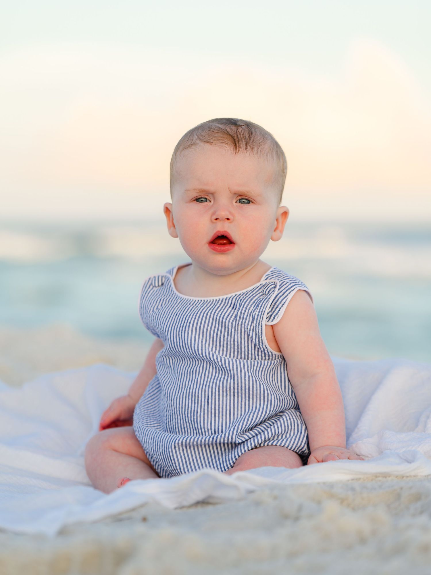 A baby in a striped romper sits on a white blanket at the beach with ocean waves in the background.