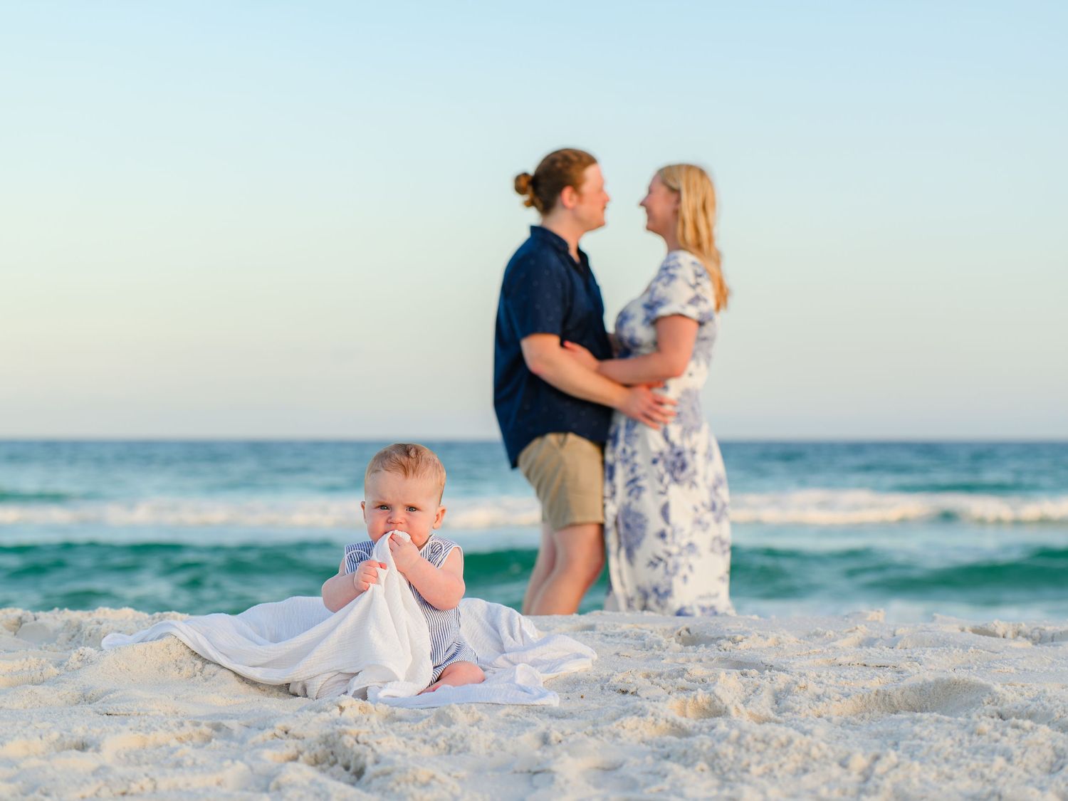 Family beach portrait with couple embracing and toddler sitting on white sand with ocean backdrop at sunset.