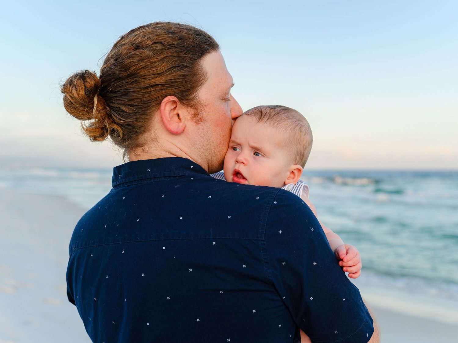 A parent kissing a baby on the cheek while holding them on a serene beach at sunset.
