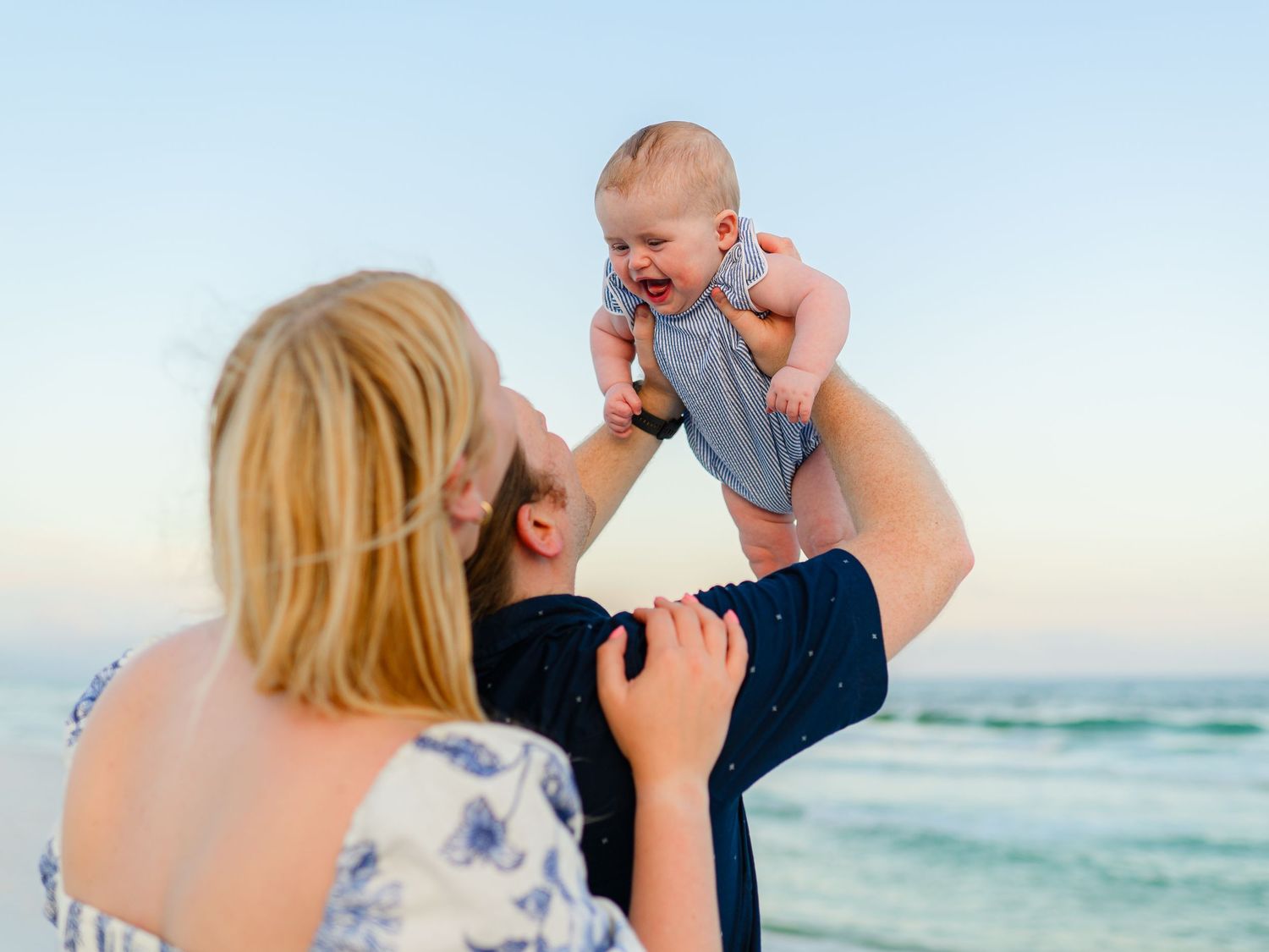 A happy family at the beach lifts their laughing baby in the air at sunset near the ocean.