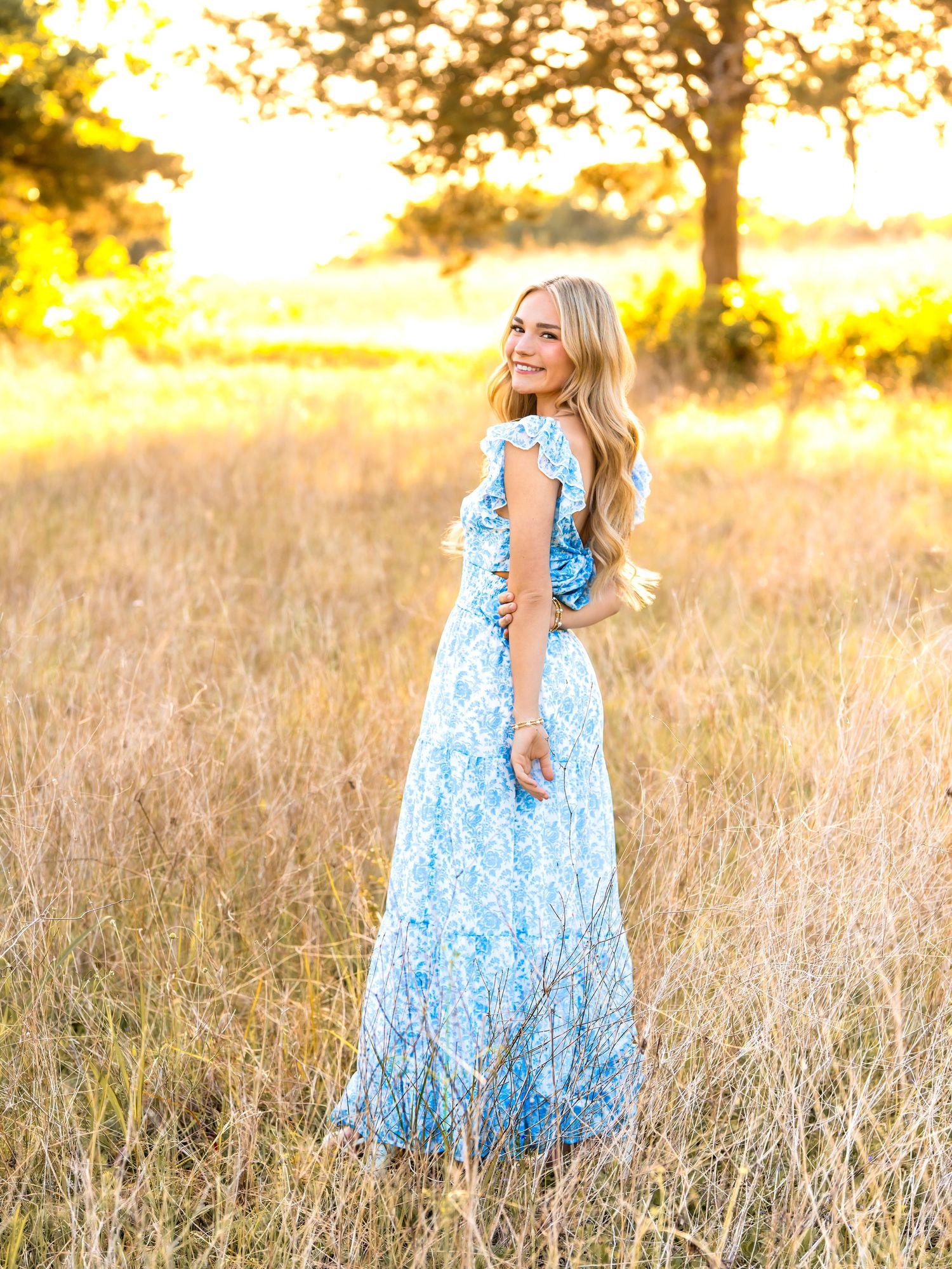Young woman in blue floral maxi dress smiling in golden field at sunset with trees in background.