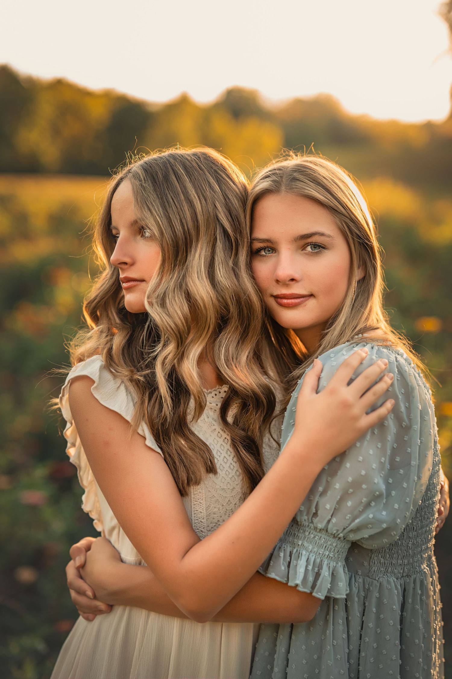 Two teenage sisters with long blonde hair embrace in a golden sunset field, wearing elegant white and blue dresses.