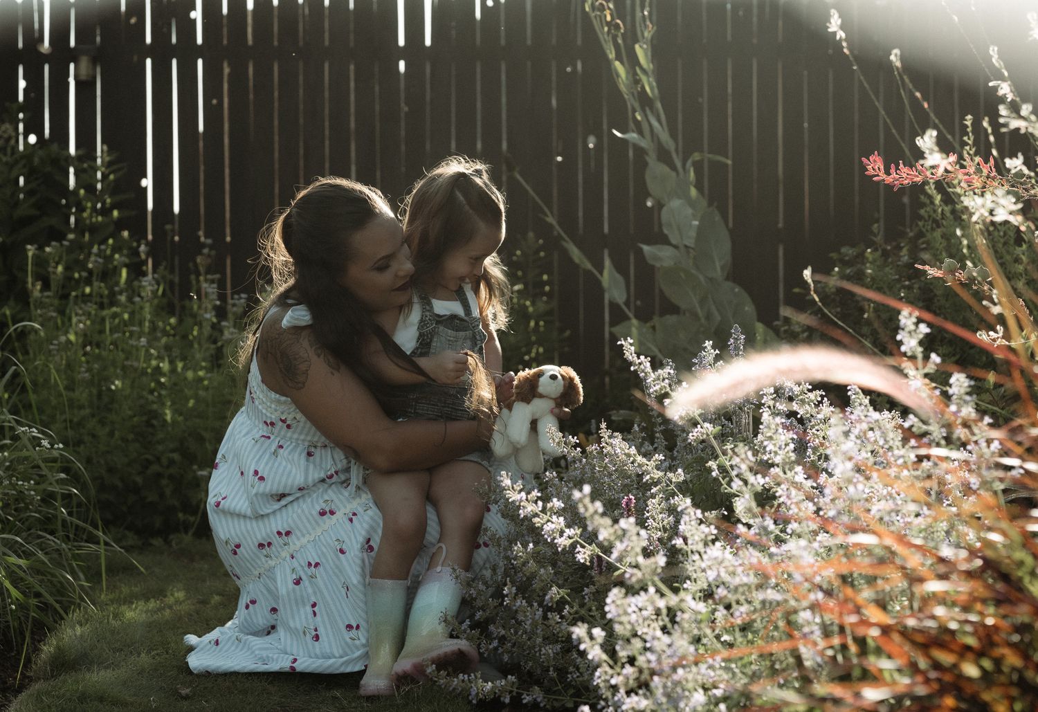 A mother and young daughter kneel together in a sunlit garden, gently petting a small rabbit among wildflowers.