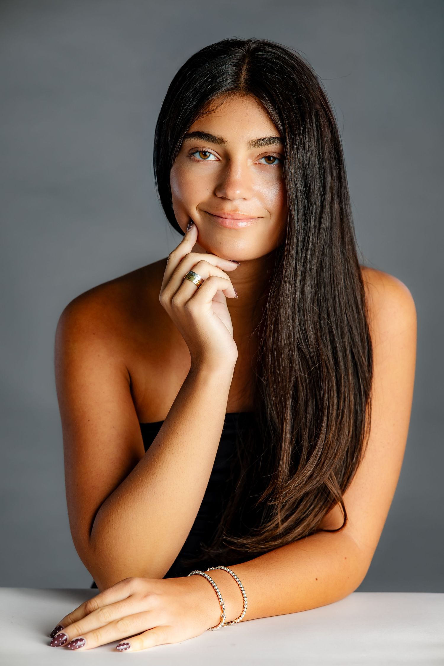 Young woman with long dark hair poses thoughtfully at a white table, wearing a black top and gold bracelet.