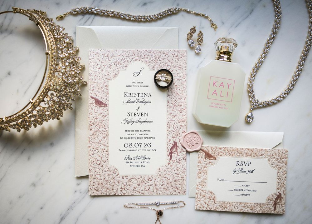Elegant wedding invitation suite with blush floral details, wax seal, perfume bottle, and jewelry on marble.
