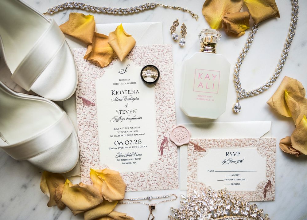 Elegant wedding invitation suite with blush floral details, RSVP card, rings, perfume, jewelry, and satin heels on marble.