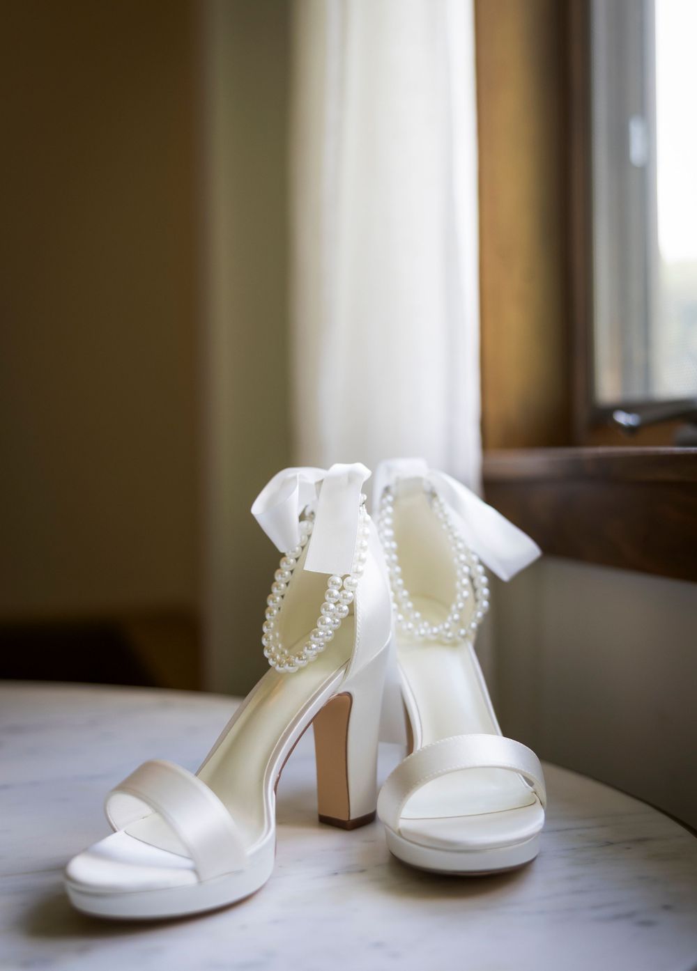 Elegant white bridal heels with pearl ankle straps and satin bow displayed on a marble surface.