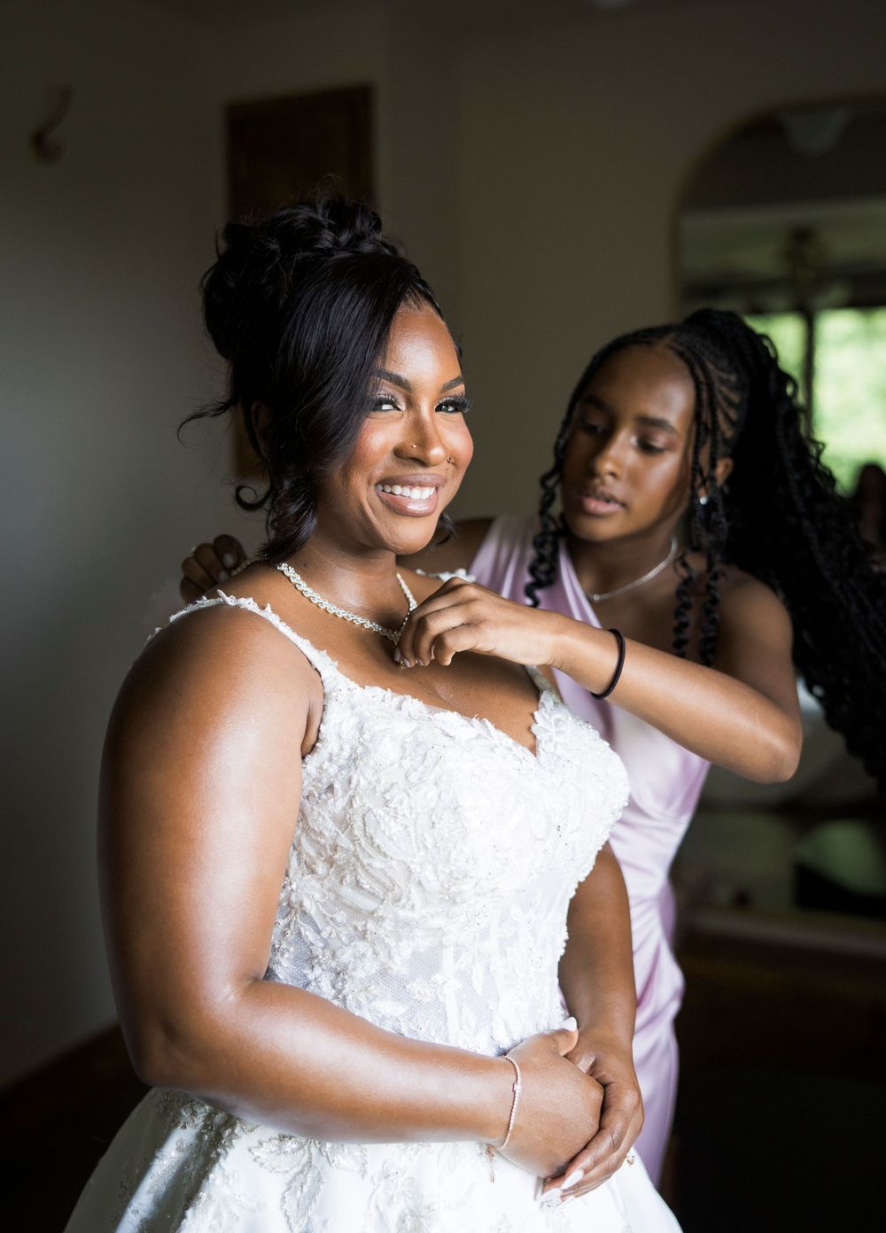 A bride in a white lace gown smiles as her bridesmaid fastens her necklace during wedding preparations.