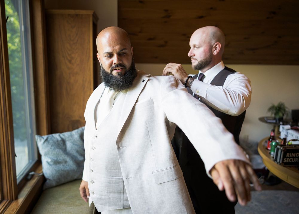 Groomsman helping bald bearded groom in white linen suit adjust his jacket before wedding ceremony.