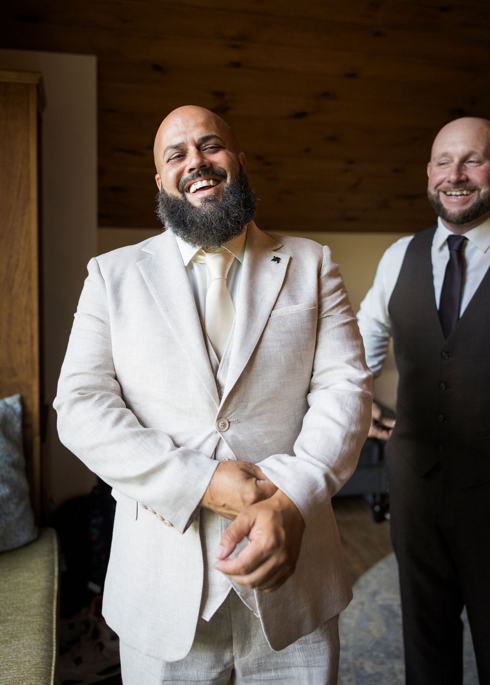 Smiling bald groom with beard in white suit laughing with groomsman in black vest behind him.