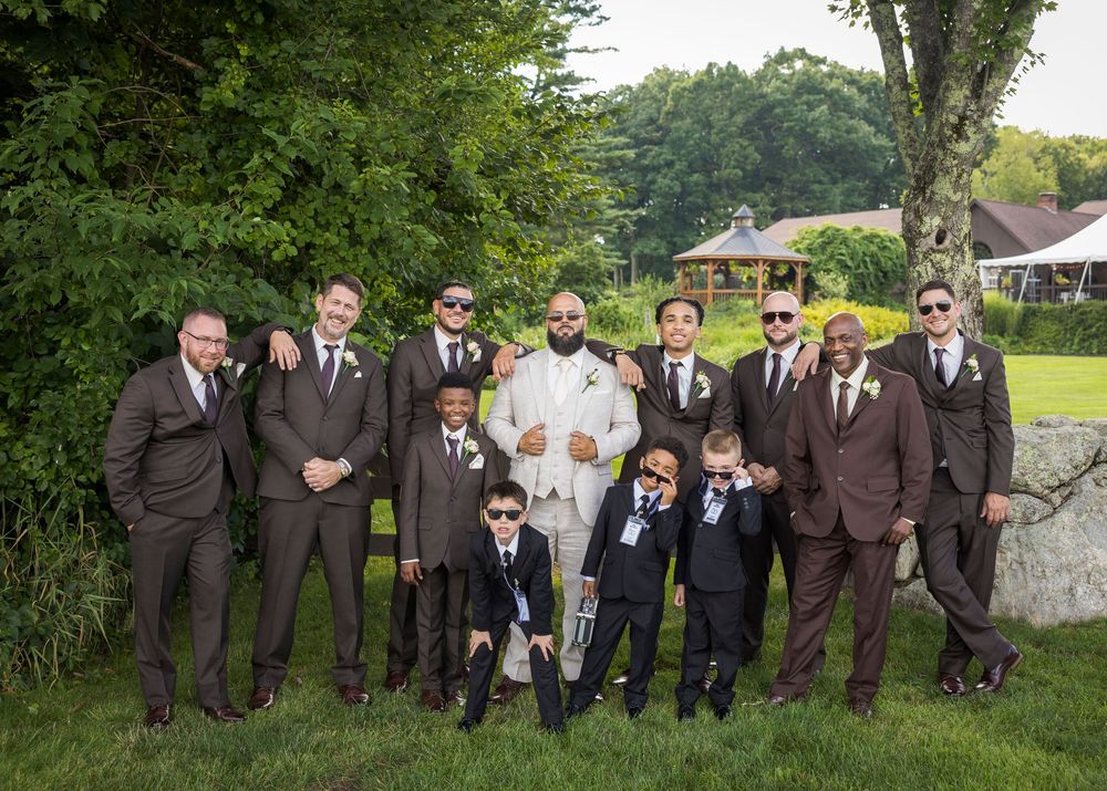 Groom in white suit poses with groomsmen and ring bearers in brown suits outdoors at a wedding.