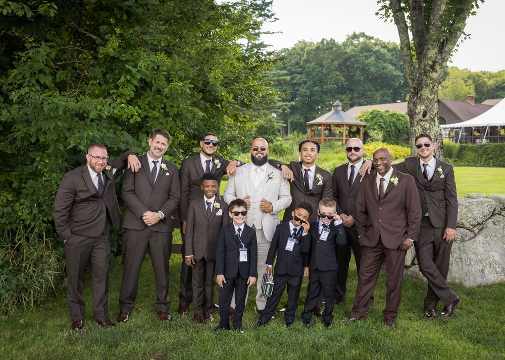 Groom in white suit poses with groomsmen and ring bearers in brown suits outdoors at a wedding venue.