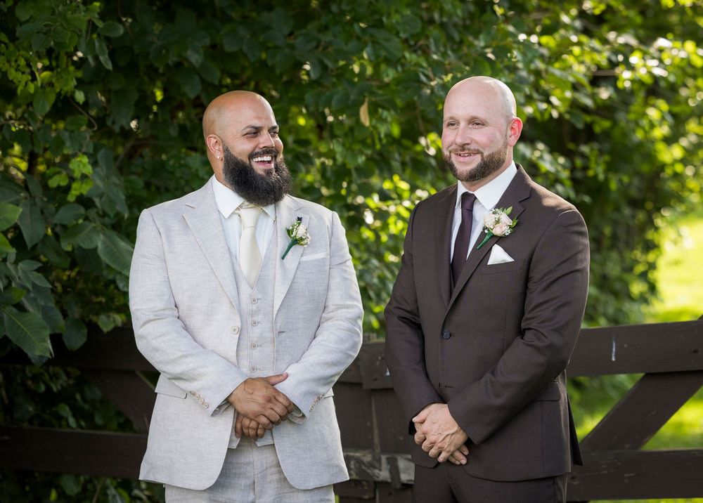 Two grooms smile outdoors in suits with boutonnieres at a garden wedding ceremony.