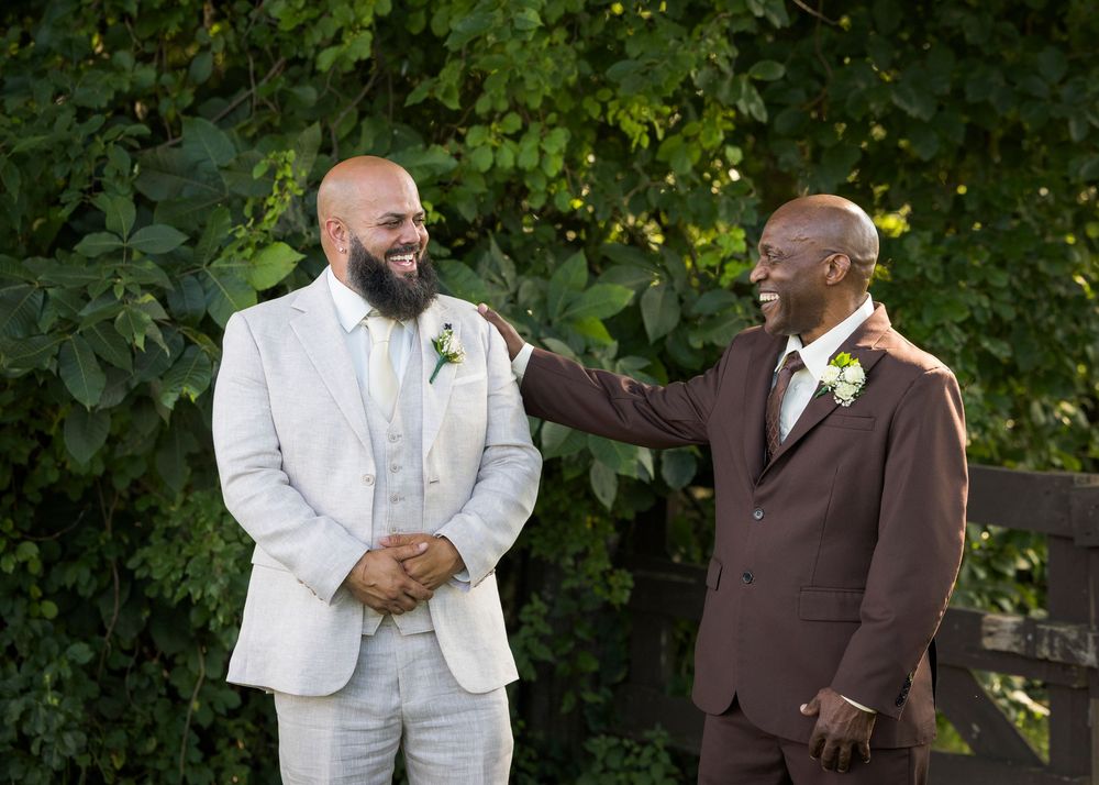 Groom in white suit shares a joyful moment with older man in brown suit at outdoor wedding.