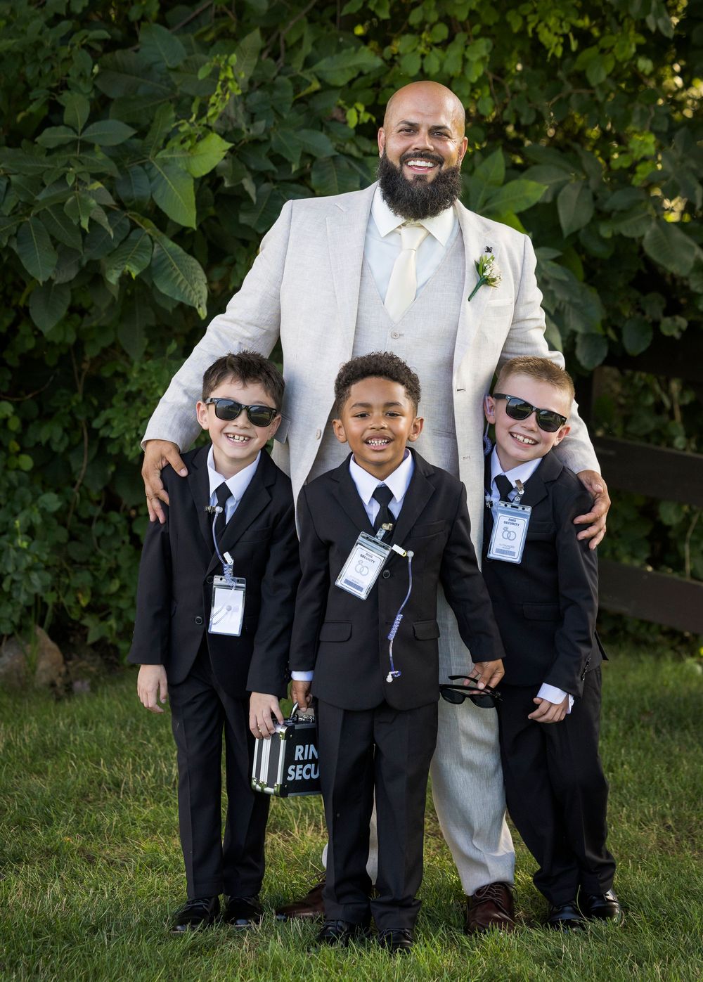 Groom in ivory suit poses with three young ring bearers wearing black suits and sunglasses at outdoor wedding.