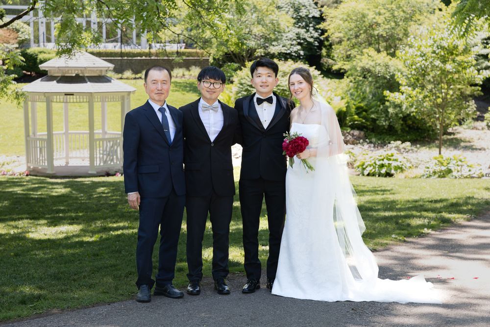 Bride and groom stand with groom father and brother surrounded by greenery at the civic garden complex in london, ontario.